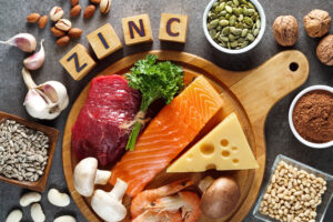 A flatlay of some of the best food sources of zinc on a round wooden serving board against a grey background. Zinc food sources of animal origin products such as meat, fish, cheese and eggs are laid out alongside vegan and plant-based sources such as pumpkin seeds, legumes, mushrooms and a variety of nuts.
