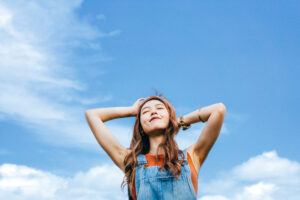 Vitamin B12 benefits blog image of a beautiful young Asian woman with eyes closed open arms and taking deep breath outdoors in nature, against blue sky on a sunny day. Enjoying sunshine and freedom in nature