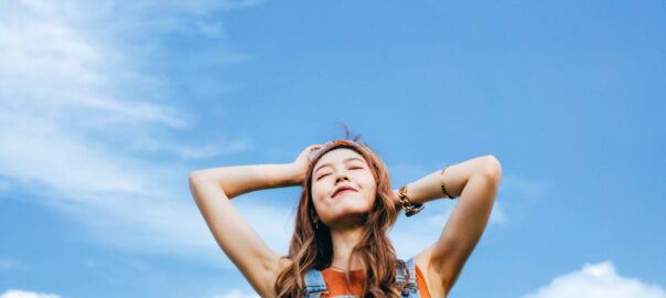 Vitamin B12 benefits blog image of a beautiful young Asian woman with eyes closed open arms and taking deep breath outdoors in nature, against blue sky on a sunny day. Enjoying sunshine and freedom in nature