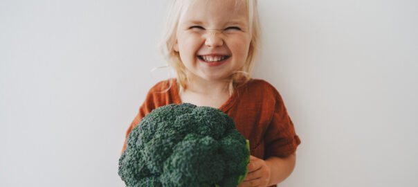 Healthy eating for children: blonde haired toddler girl with a cheeky grin holding broccoli.