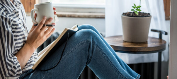 Young woman sat holding a mug whilst journalling. Journalling may help when trying to build and reinforce healthful habits.