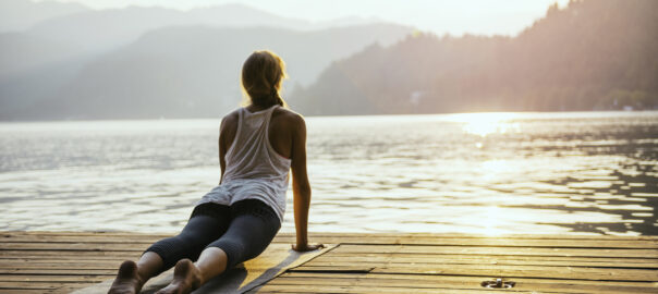 Woman doing yoga by a lake at sunrise. She is in upward dog pose and wears a vest and leggings. Yoga can help to balance stress and cortisol levels.