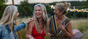 Three white women over 50 laughing together in a field at a festival with fairy lights in the background.