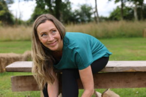 Smiling mid-life woman with long hair sits on an outdoor wooden bench by a lake to tie her shoe laces before going for a run. She wears a teal t-shirt and black leggings.