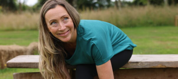 Smiling mid-life woman with long hair sits on an outdoor wooden bench by a lake to tie her shoe laces before going for a run. She wears a teal t-shirt and black leggings.